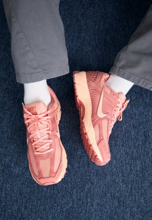 Stopy w różowych butach sportowych Nike, białych skarpetkach i szarych spodniach stojące na ciemnoniebieskim dywanie.