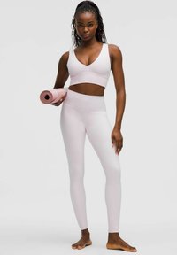 Svijetlo ružičasti sportski crop top i tajice izrađeni od rastezljivog materijala, s glatkom teksturom. Model drži smotanu jogu prostirku.