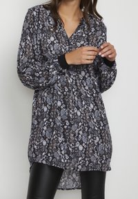 Tunique imprimée florale en noir, avec manches longues, décolleté en V et poignets côtelés noirs. Tissu léger avec une coupe ample.