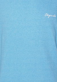 Ljusblå T-shirt med ett texturerat mönster av små vita prickar. Har "Originals" broderat i vit tråd på framsidan.