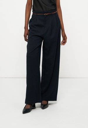 Άτομο που φοράει μαύρο παντελόνι ψηλόμεσο, με φαρδύ πόδι και λεπτή καφέ ζώνη, καθώς και μαύρες μυτερές γόβες, σε λευκό φόντο.