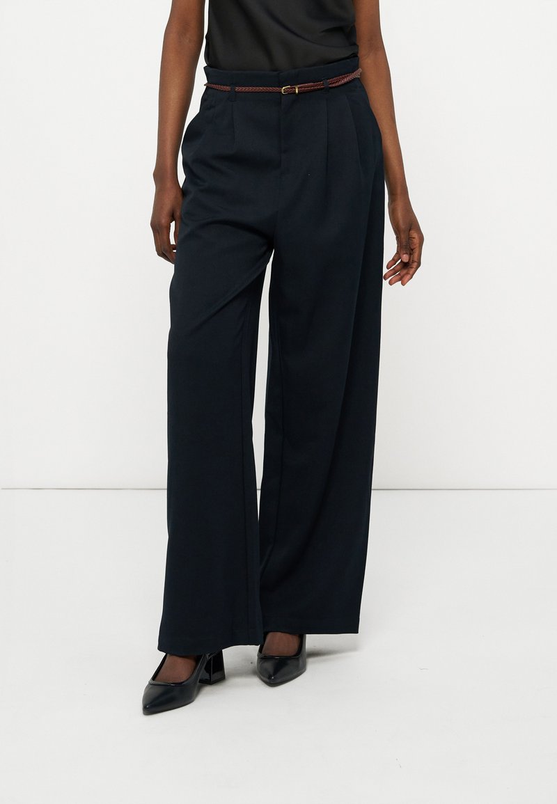 Άτομο που φοράει μαύρο παντελόνι ψηλόμεσο, με φαρδύ πόδι και λεπτή καφέ ζώνη, καθώς και μαύρες μυτερές γόβες, σε λευκό φόντο.