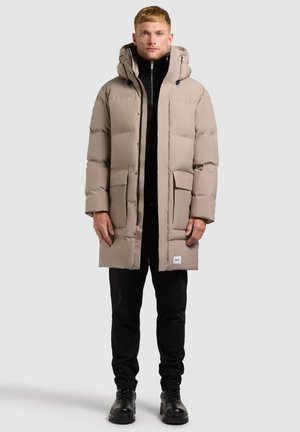 Chaqueta parka de color beige con capucha, con bolsillos amplios, cremallera frontal y una etiqueta de marca sutil cerca del dobladillo. Llevada sobre pantalones negros.