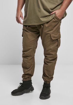 Mann trägt olivgrüne Cargohose mit elastischen Bündchen und schwarze Sportschuhe, steht mit einer Hand in der Tasche vor grauem Hintergrund.