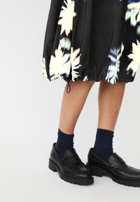 Czarna kwiecista spódnica midi z marszczonym dołem, zestawiona z czarnymi lordami i granatowymi skarpetkami. Spódnica ma duże wzory kwiatowe i ozdobny sznurek.