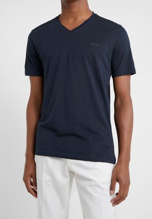 Homme portant un T-shirt à col en V bleu marine avec le logo "BOSS" sur la poitrine et un pantalon blanc, debout devant un fond uni.