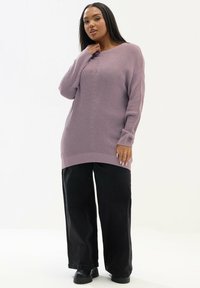 Lila gestrickter Pullover mit langen Ärmeln und einem gerippten Muster, kombiniert mit weiten schwarzen Hosen und klobigen schwarzen Stiefeln.