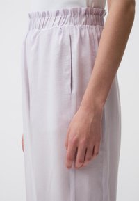 Helle Lavendelhosen mit hoher Taille, gerafftem elastischen Bund, seitlichen Taschen und glatter Textur, abgebildet mit einer Hand am Saum.