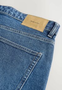 Denim bikses ar gaiši zilu nokrāsu, ar brūnu ādas etiķeti ar uzrakstu "DENIM SUPPLY" pie jostas un klasiskām šuvju detaļām.
