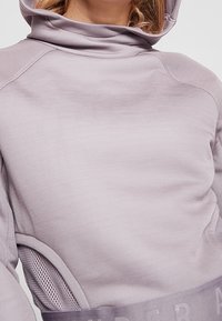 Vaaleanlila huppari, jossa on korkea kaulus, verkkohuokosia ja leveä vyötärönauha, jossa on teksti. Sileä kankaankuvio, rento istuvuus.