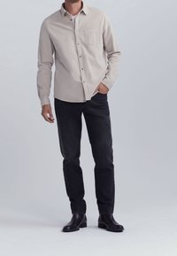 Hombre vestido con una camisa de botones de manga larga beige, jeans negros y botas de cuero negras, de pie con una mano en el bolsillo sobre un fondo plano.