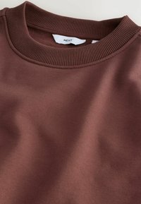Hanorac maro cu guler rotund fabricat dintr-un material neted, cu o linie de guler cu nervuri. Prezintă detalii de cusătură și etichetă. Suprafață texturată vizibilă.