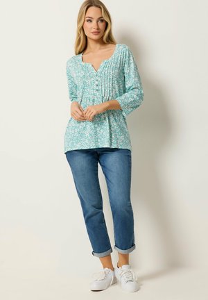 Frau steht vor einer Wand, trägt eine hellblaue Blumenbluse mit Knöpfen und Falten, blaue hochgekrempelte Jeans und weiße Turnschuhe.