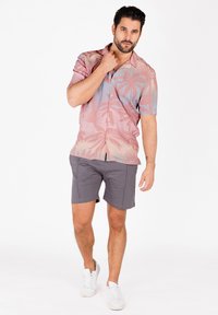 Kurzarmhemd in Rosa und Blau mit Palmenblattmuster, kombiniert mit grauen Shorts. Das Modell trägt weiße Turnschuhe und hat die Hände am Kragen.