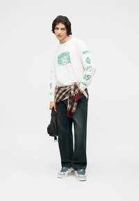 Νέος άνδρας με άσπρο μακρυμάνικο πουκάμισο με γραφικά, πουλόβερ καρό δεμένο στη μέση, σκουρόχρωμο τζιν, λευκά αθλητικά παπούτσια και κρατώντας μια μικρή μαύρη τσάντα.