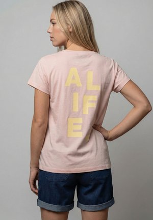 Frau mit langen blonden Haaren, die ein hellrosa T-Shirt mit großen gelben Buchstaben "ALIFE" auf dem Rücken und dunkelblaue, hochgekrempelte Shorts trägt.