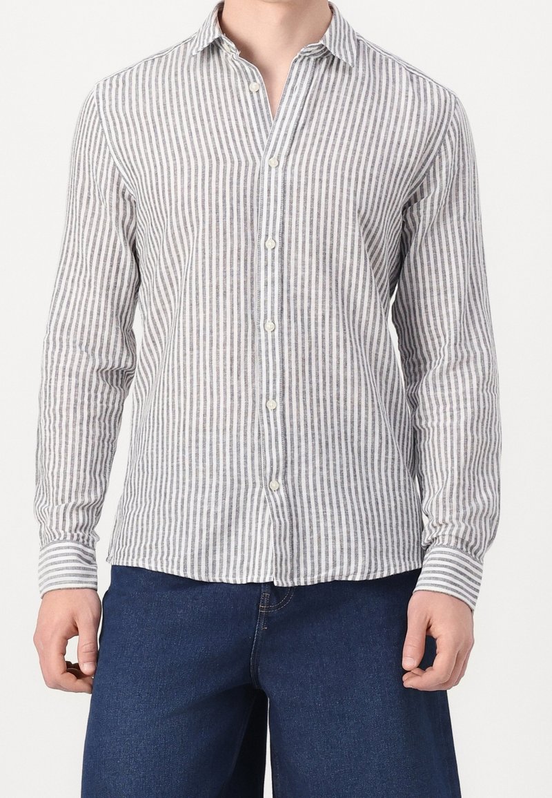 Homme portant une chemise blanche à manches longues avec de fines rayures verticales grises, associée à un jean bleu foncé.