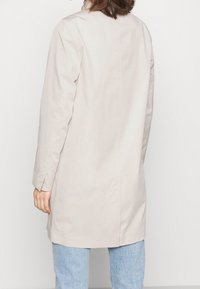 Ljusbeige kappa med rak skärning, långa ärmar och en ren bakseam. Tyget har en slät textur och minimal detaljer.