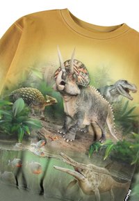 Mikina s dlhým rukávom v zlatej farbe s potlačou dinosaura. Obsahuje textúrované detaily, živé farby a obrázky s džungľovým pozadím.