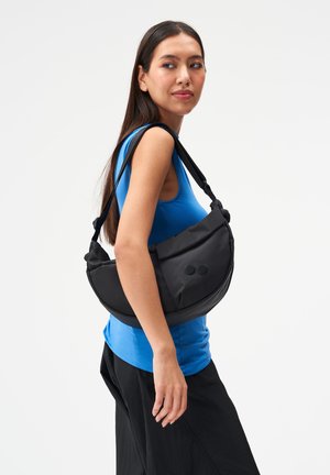 Frau trägt ein blaues ärmelloses Oberteil und schwarze Hose und hält eine große schwarze taschenförmige Schultertasche mit zwei runden Knöpfen.