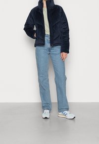 Granatowa kurtka sztruksowa z wysokim kołnierzem, zamkiem błyskawicznym i pikowanymi detalami, zestawiona z jasnoniebieskimi jeansami o prostych nogawkach oraz szarymi tenisówkami.