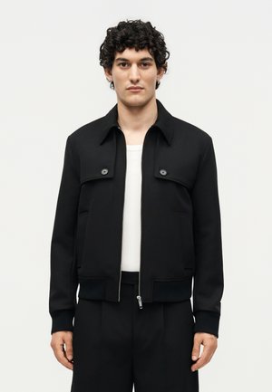 Άνδρας με κοντά σγουρά μαλλιά που φοράει μαύρο μπουφάν με φερμουάρ πάνω από λευκό πουκάμισο, στέκεται μπροστά σε ένα απλό ανοιχτό φόντο.