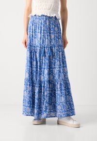 Blaue Maxiröhre mit weißen Mustern, gestuftem Design, elastischem Bund und Quastenakzenten. Kombiniert mit weißen Sneakers.