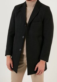 Cappotto in misto lana nero con design sartoriale, doppiopetto con bottoni, rever con intaglio, tasche laterali e taschino sul petto, indossato sopra un maglione beige a maglia.
