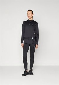 Haut de sport noir à manches longues avec fermeture éclair au quart et logo, associé à des leggings noirs ajustés et une ceinture noire avec porte-bouteille d'eau.