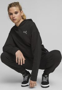 Čierna mikina s kapucňou a bielym logom Puma, v kombinácii s rovnako farebnými teplákmi a čiernymi platformovými teniskami s bielymi akcentmi a textúrovanými šnúrkami.