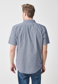 Krátký rukáv košile v modro-bílé geometrické vzoru, s špičatým límcem a zaobleným dolním okrajem, vyrobená z lehké tkaniny.