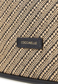 Borsa in paglia intrecciata con un motivo zigzag nero e beige, con un'etichetta in pelle testurizzata impressa con "COCCINELLE" in lettere dorate.