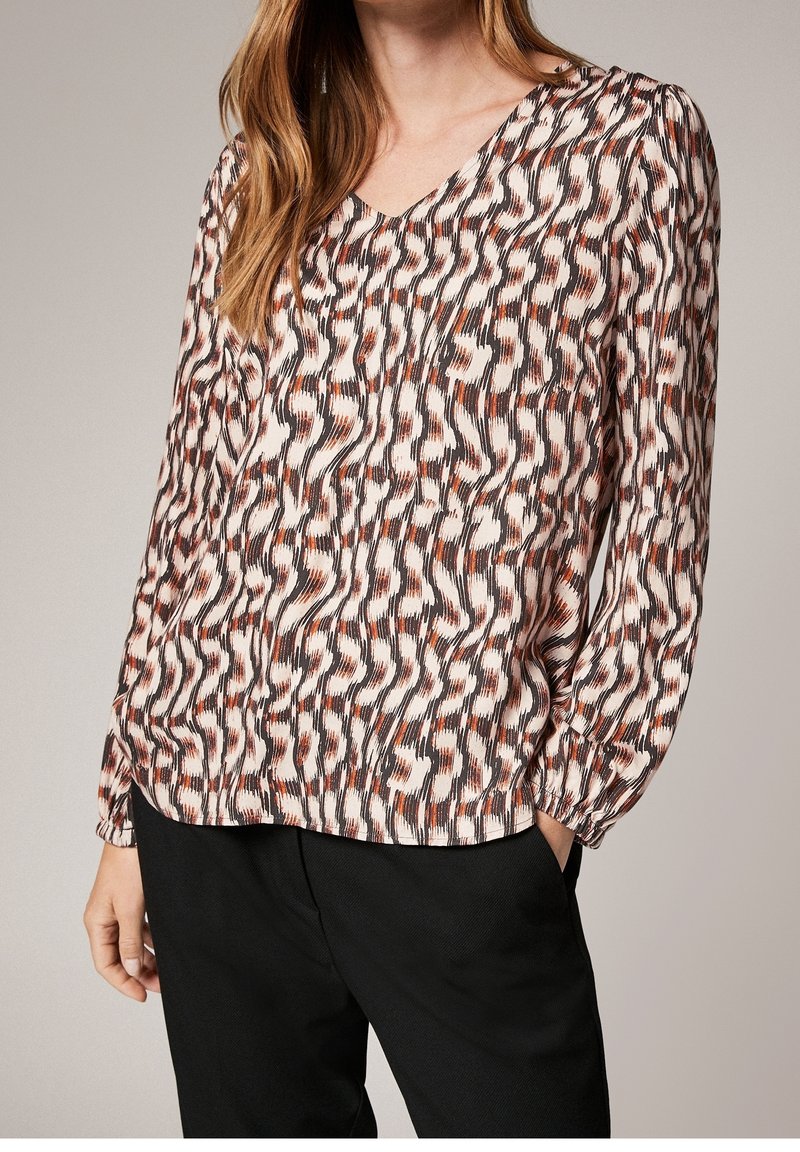 Femme portant un chemisier à manches longues à motif abstrait marron, noir et beige, associé à un pantalon noir, debout avec une main dans la poche.