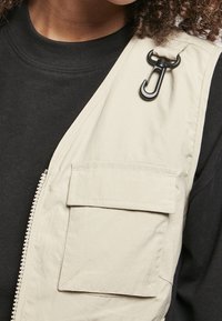 Beżowa kamizelka bez rękawów wykonana z lekkiego materiału, z przednią kieszenią i czarnym akcentem z metalowego zapięcia. Noszona na czarnej koszuli.