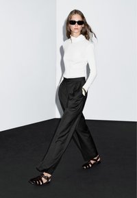 Witte, nauwsluitende, lange mouwen top, geplooide zwarte broek en zwarte sandalen met bandjes. Glad materiaal, strakke vormgeving, met een minimalistische esthetiek.