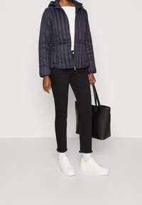 Marineblau gepolsterte Jacke mit Kapuze, Reißverschluss vorne und gestepptem Muster, kombiniert mit schwarzen Hosen und weißen Sneakern sowie einer schwarzen Tragetasche.