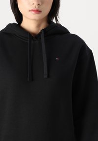 Felpa nera con cappuccio realizzata in materiale morbido. Presenta una tasca frontale, lacci e un piccolo logo sul lato sinistro del petto.