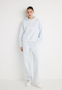 Sweatshirt com capuz azul-claro e bolso frontal, combinado com calças de fato de treino coordenadas. Modelo usa ténis brancos. Textura lisa em toda a peça.