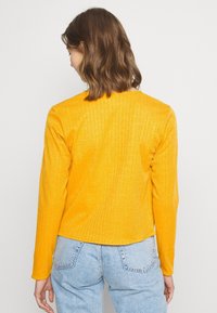 Žlutý vroubkovaný top s dlouhým rukávem a kulatým výstřihem, navržený v přiléhavém střihu, v kombinaci se světlými modrými džínovými kalhotami.