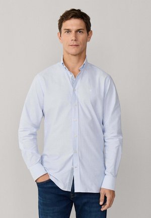 Homme portant une chemise à manches longues bleu clair boutonnée et un jean bleu foncé, debout avec une main dans la poche devant un fond uni.