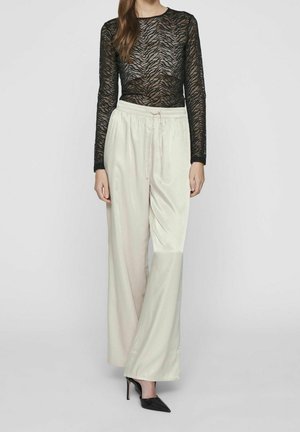 Žena v černém průsvitném topu s dlouhým rukávem a motivem tygra, krémových saténových kalhotách širokých nohavic s šňůrkou a černých špičatých vysokých podpatcích.