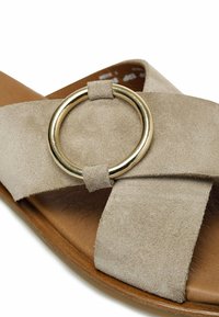 Sandalia de ante beige con correas entrecruzadas y un acento circular en tono dorado. La plantilla es marrón y tiene una textura suave.