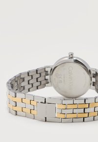 Μεταλλικό λουράκι ρολογιού με ασημί και χρυσούς κρίκους, συνδεδεμένο σε στρογγυλή θήκη ρολογιού Calvin Klein που δείχνει την πίσω πλευρά.