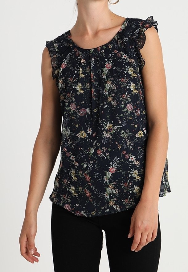 Blouse noire sans manches avec imprimé floral multicolore et bordure froncée aux épaules, portée avec un pantalon noir.