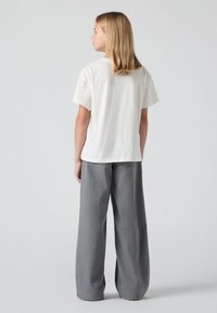 Jaunas žmogus su pečių ilgio blondiniais plaukais, apsirengęs balta trumpomis rankovėmis marškiniu ir plačiomis pilkomis kelnėmis, atsisukęs į ganėtinai neutralų foną.