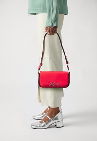 Borsa a tracolla rossa in pelle con chiusura a pattina, accessori in argento e lettera "Kvl", indossata con pantaloni chiari e tacchi metallici argentati.