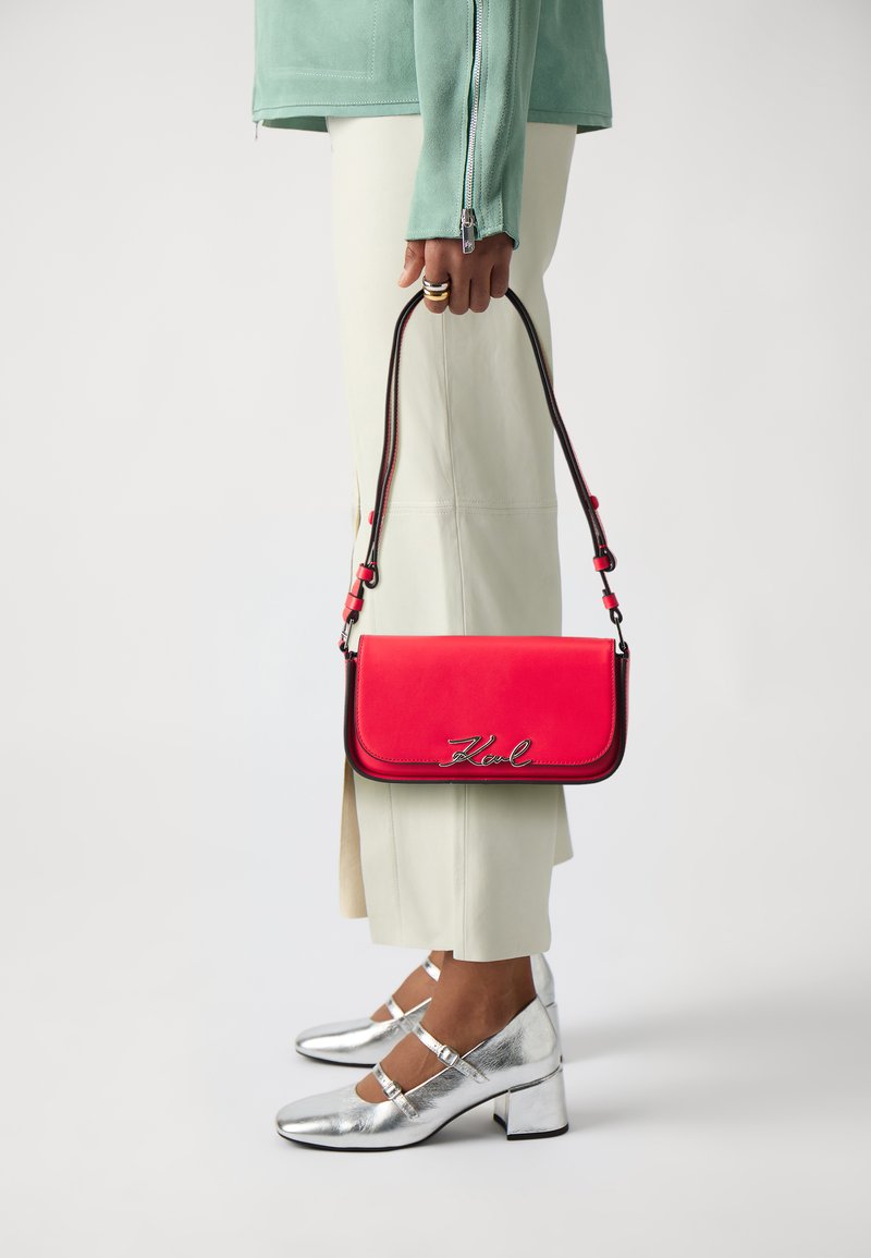 Borsa a tracolla rossa in pelle con chiusura a pattina, accessori in argento e lettera "Kvl", indossata con pantaloni chiari e tacchi metallici argentati.