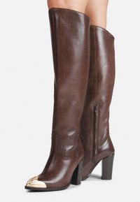 Botas de cuero marrón hasta la rodilla con un acabado brillante, que cuentan con cierre de cremallera, tacón alto y cuadrado, y una puntera decorativa en tono dorado.