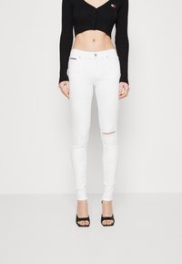 Pantalones vaqueros blancos ajustados con una rodilla desgastada, combinados con un top corto de manga larga negro con canalé. Cuenta con un cierre frontal de botón y un diseño ceñido.