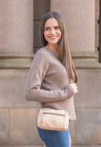 Giovane donna che sorride di lato, indossando un maglione beige, jeans blu e una piccola borsa a tracolla beige, in piedi davanti a uno sfondo di muro di pietra.